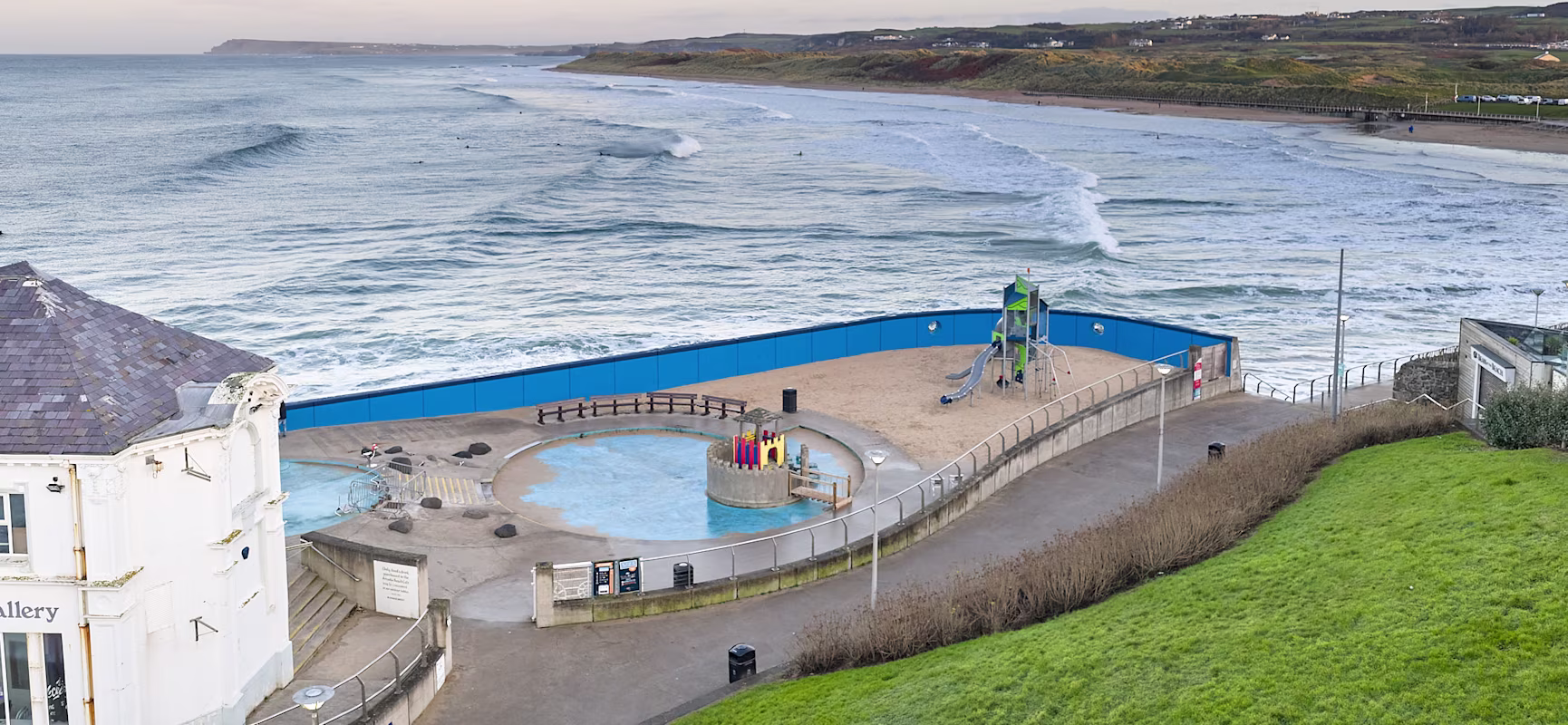 Seaside view with waves, a sandy playground and paddling pool behind blue fencing, beside a promenade and grassy slope.