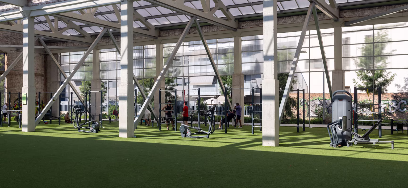 People exercising in an open-air gym under a large steel structure, with green artificial turf and fitness machines spread around.