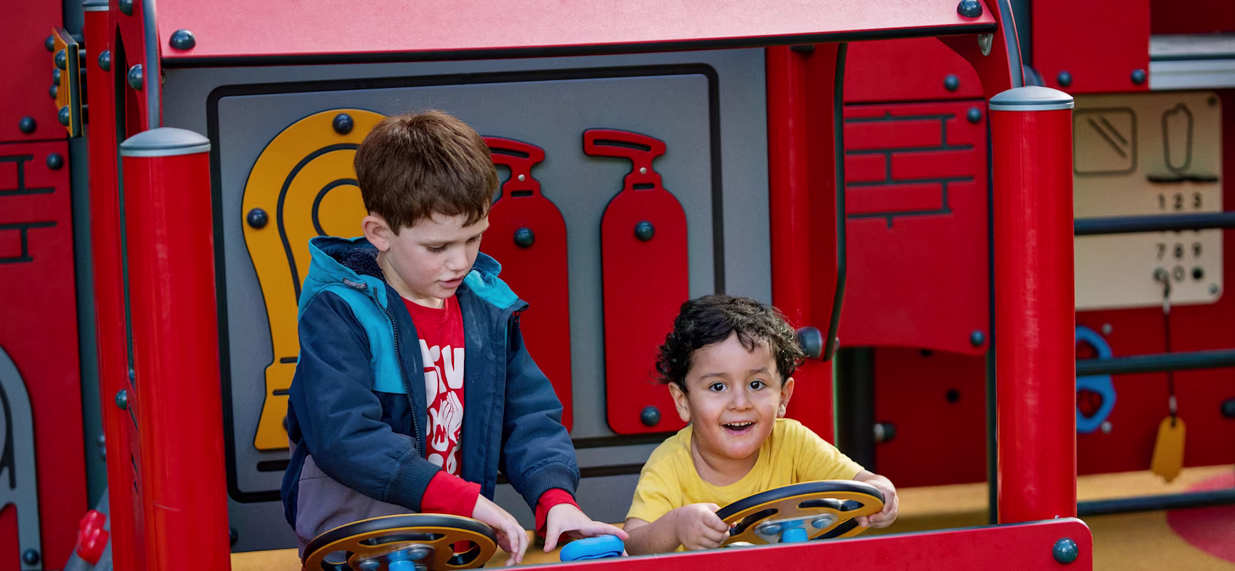 Two young children with blurred faces play in a red toy fire truck, each holding a steering wheel at an outdoor playground.