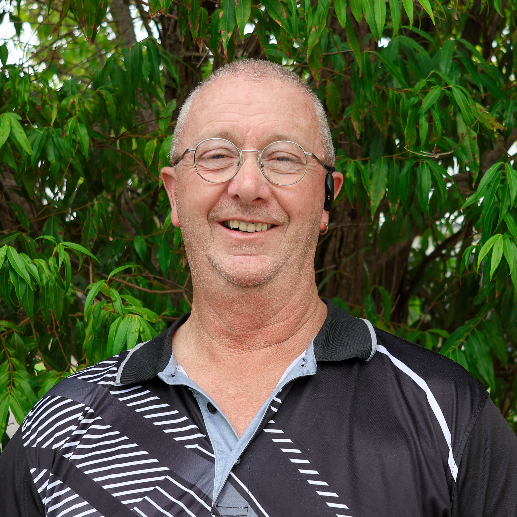 Person with blurred face wearing a black patterned polo shirt, standing outdoors in front of dense green leafy trees.