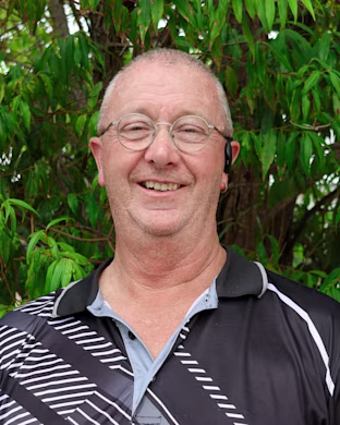 Person with blurred face wearing a black patterned polo shirt, standing outdoors in front of dense green leafy trees.