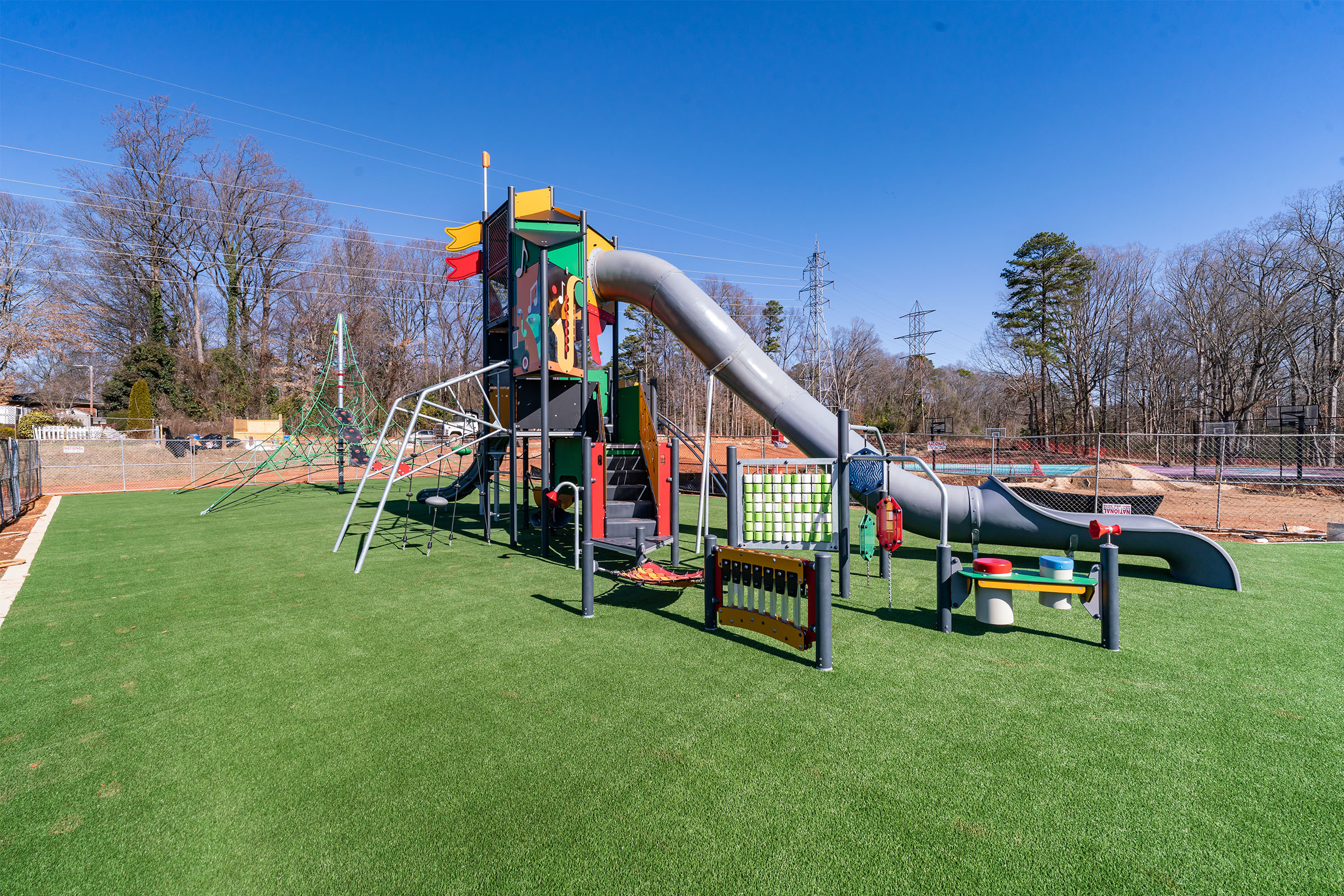 Colorful playground with tall slide, climbing nets, and activity panels on artificial grass, under a clear blue sky near trees.