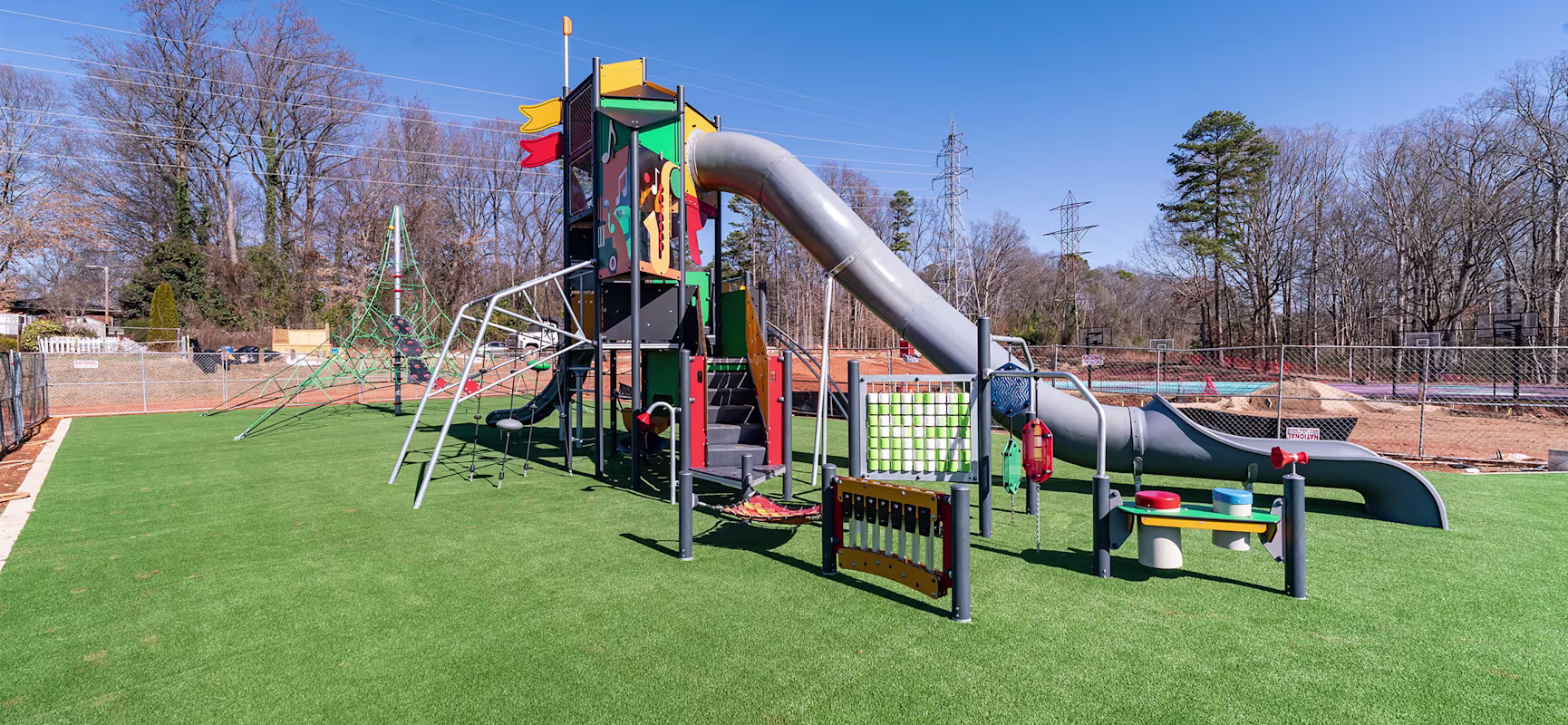 Colorful playground with tall slide, climbing nets, and activity panels on artificial grass, under a clear blue sky near trees.