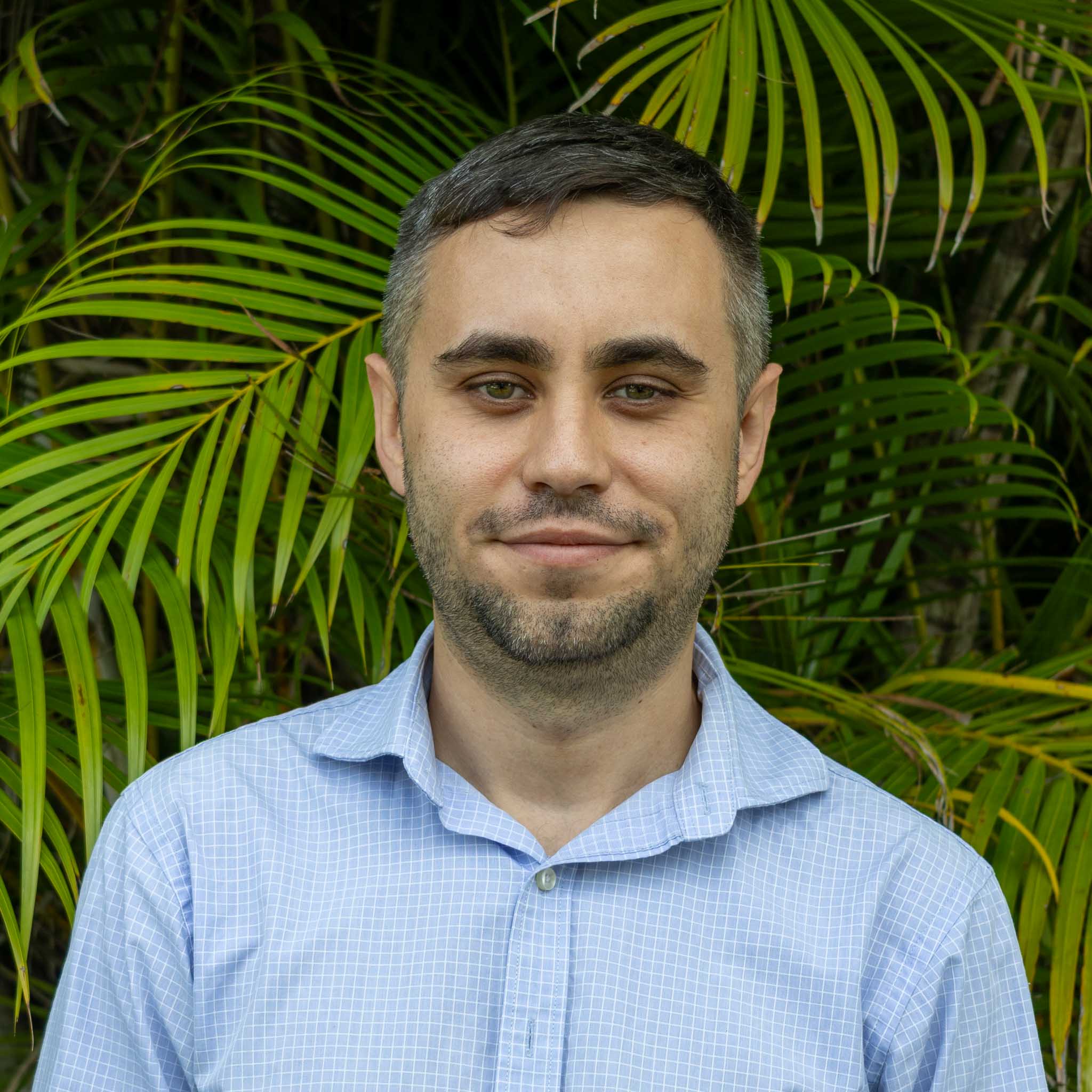 Person with blurred face wearing floral shirt and black hoodie, standing in front of lush green palm leaves outdoors.