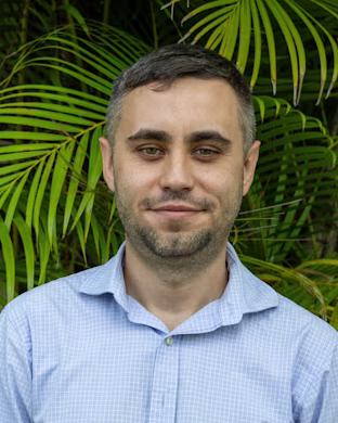 Person with blurred face wearing floral shirt and black hoodie, standing in front of lush green palm leaves outdoors.