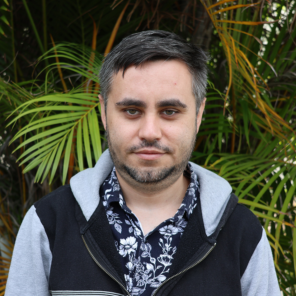 Person with blurred face wearing floral shirt and black hoodie, standing in front of lush green palm leaves outdoors.
