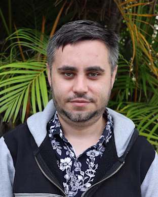 Person with blurred face wearing floral shirt and black hoodie, standing in front of lush green palm leaves outdoors.