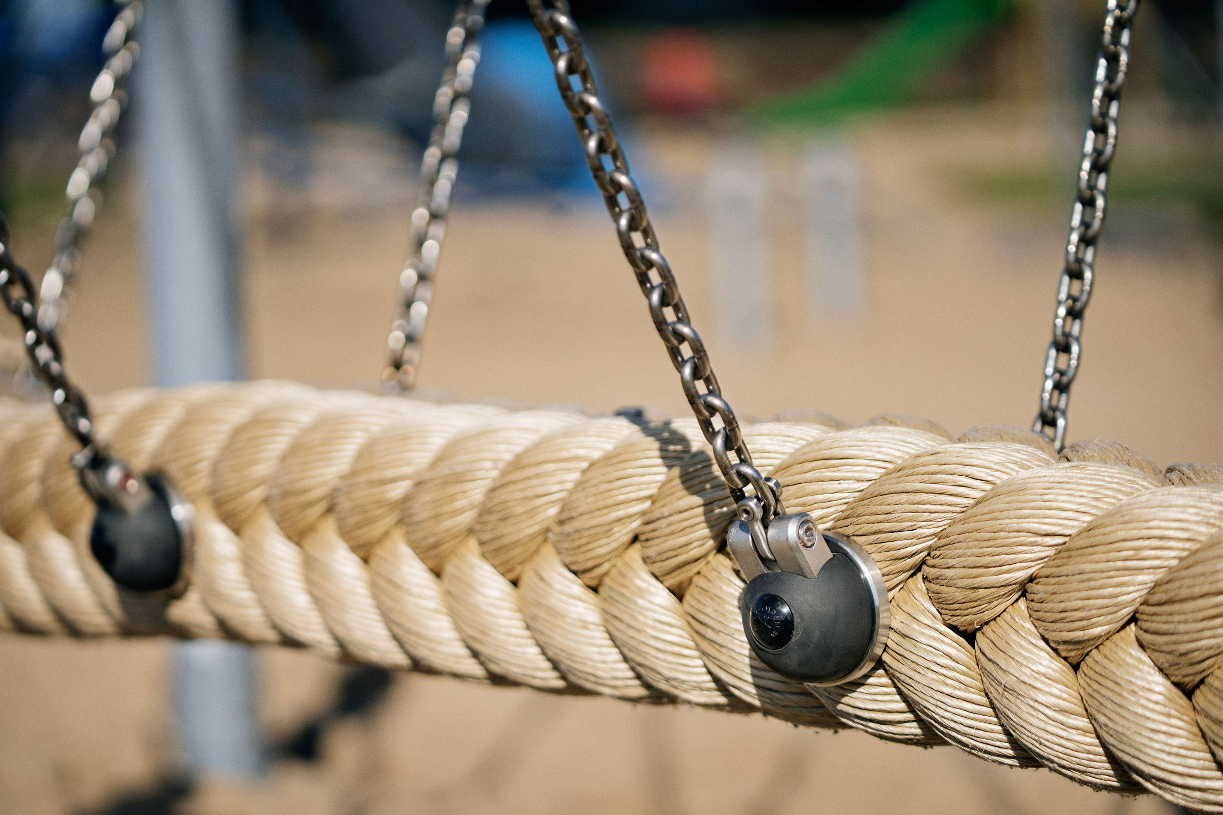 Close-up of a thick braided rope swing hanging from metal chains at a playground, with sandy ground softly blurred in the background