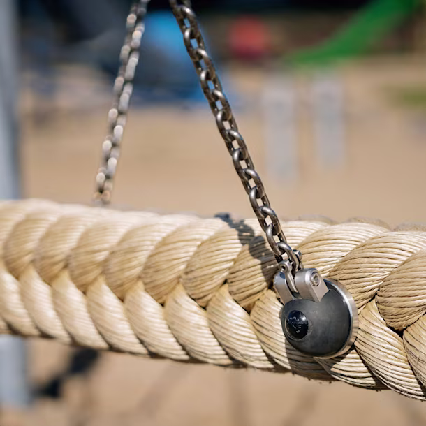 Close-up of a thick braided rope swing hanging from metal chains at a playground, with sandy ground softly blurred in the background