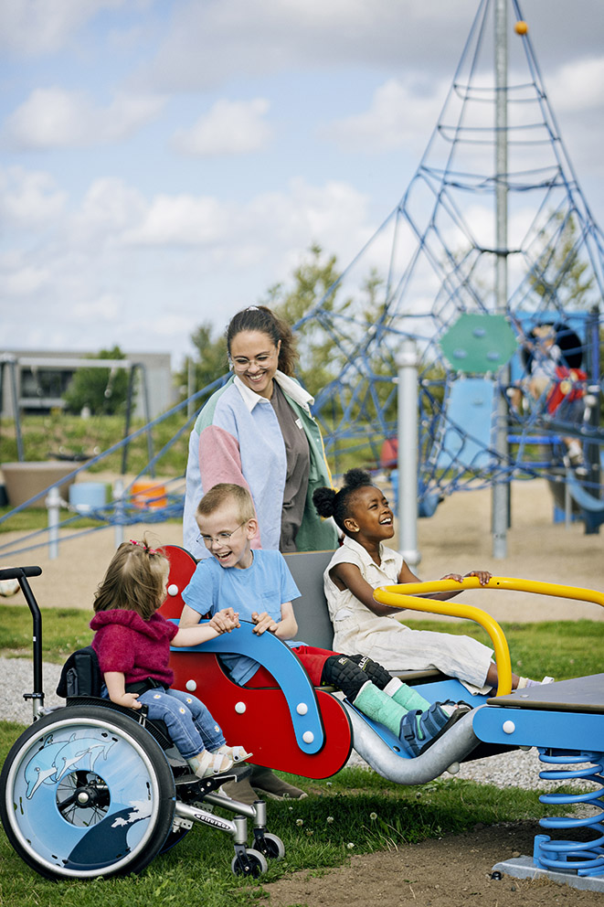 Children, including one in a wheelchair, play together on an inclusive spring rocker at a playground with climbing frames behind.