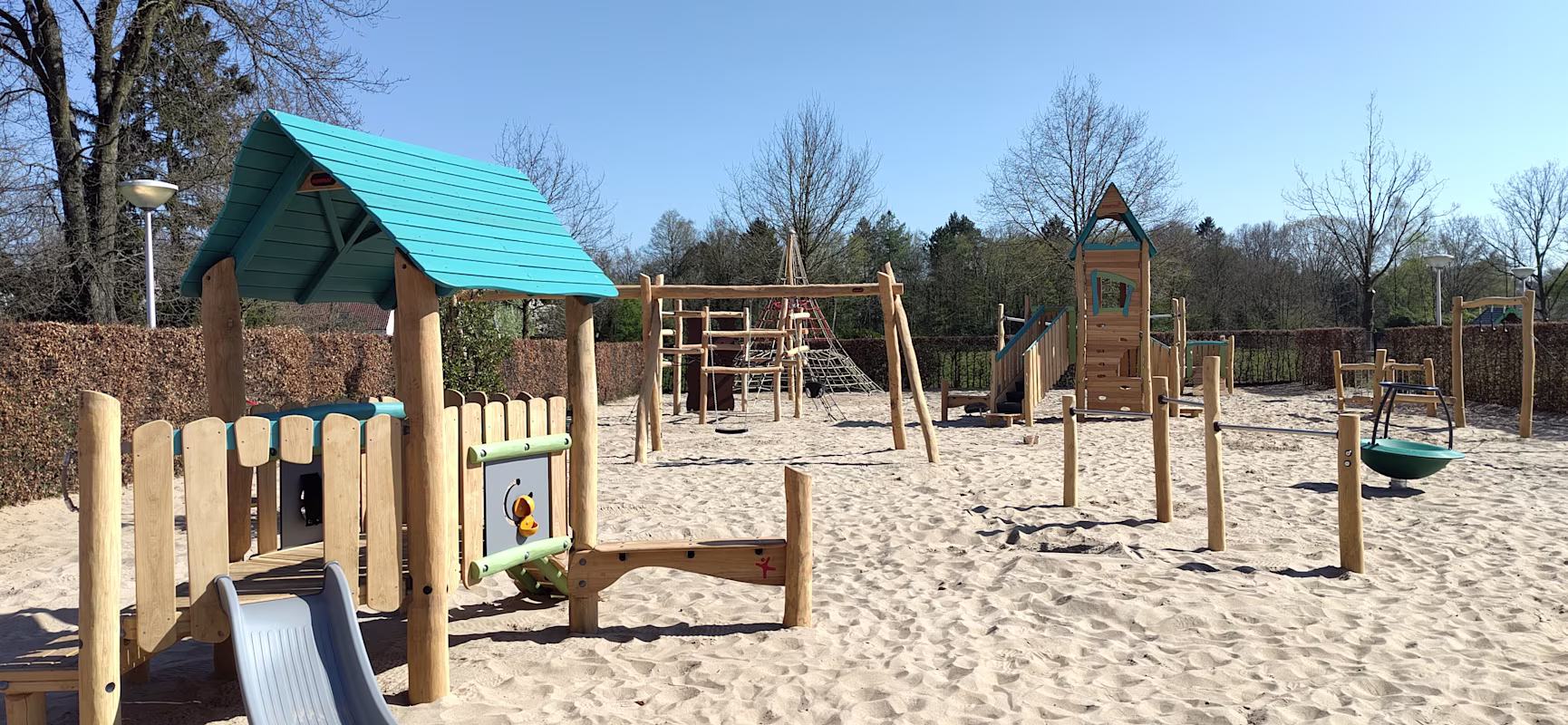 Sunny outdoor playground with wooden climbing frames, slides and swings on white sand, surrounded by trees and hedges under clear blue sky