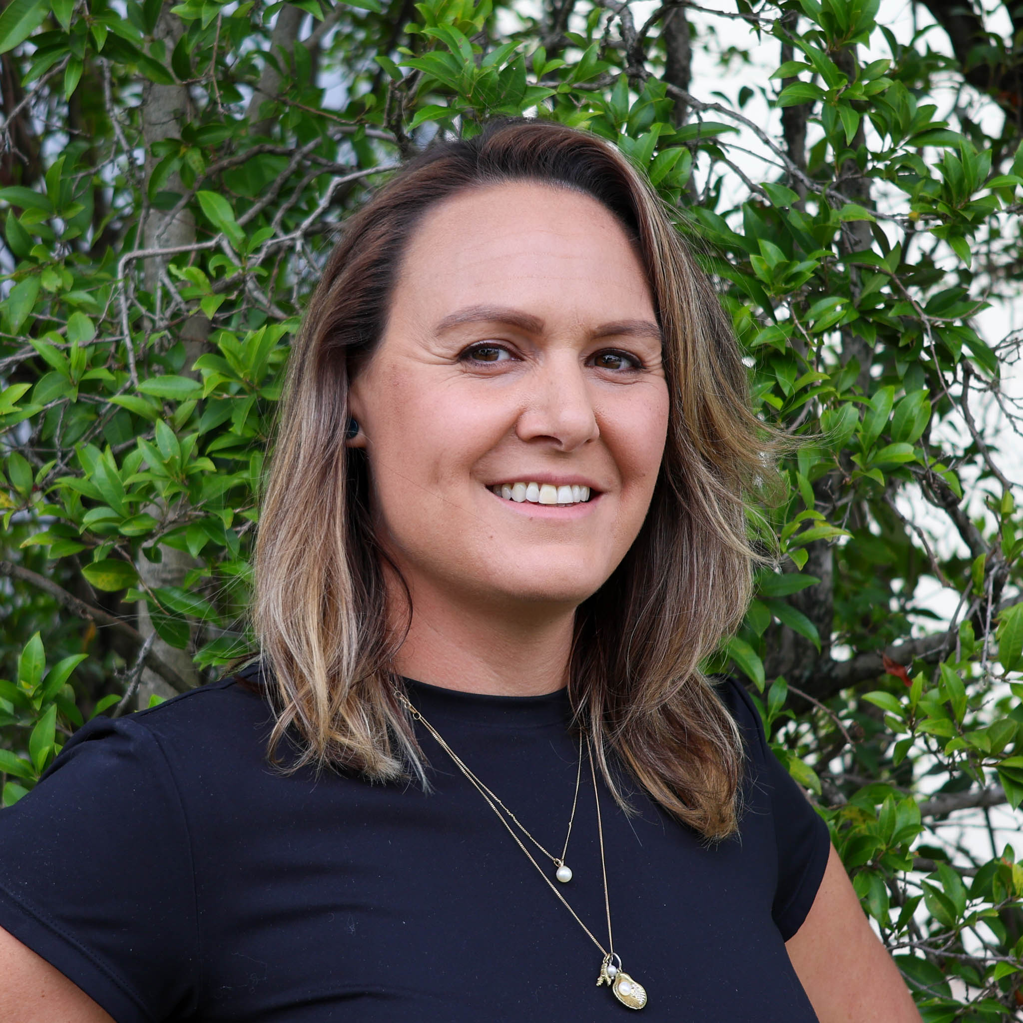 Person with blurred face in black shirt and layered necklaces standing outdoors in front of green leafy bushes