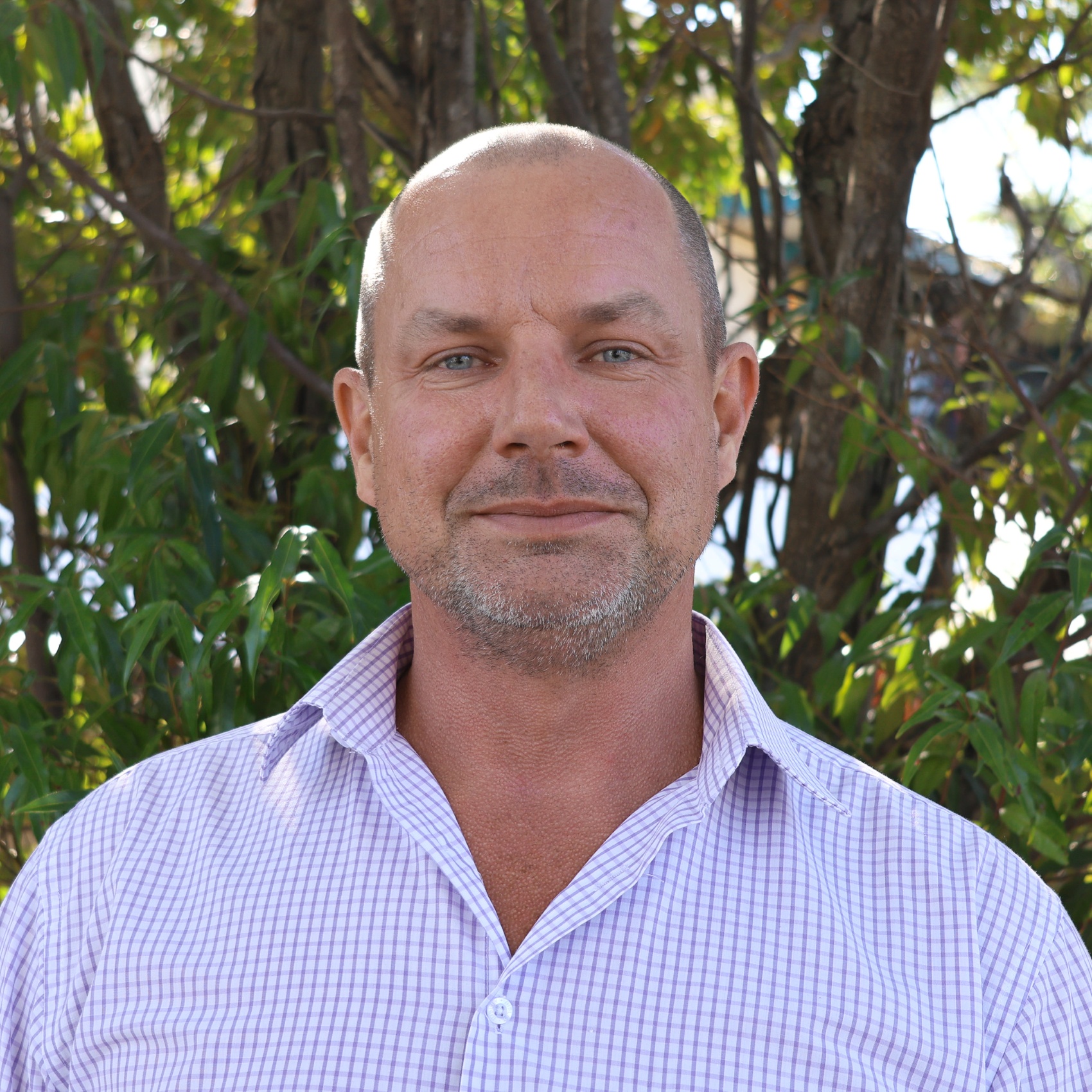 Person with blurred face in a light purple checkered shirt standing outdoors in front of leafy green trees