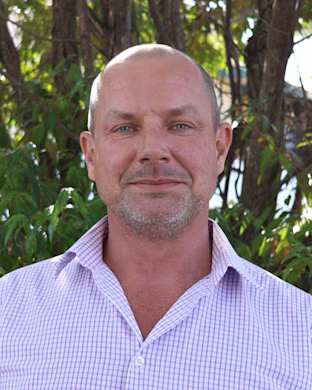 Person with blurred face in a light purple checkered shirt standing outdoors in front of leafy green trees