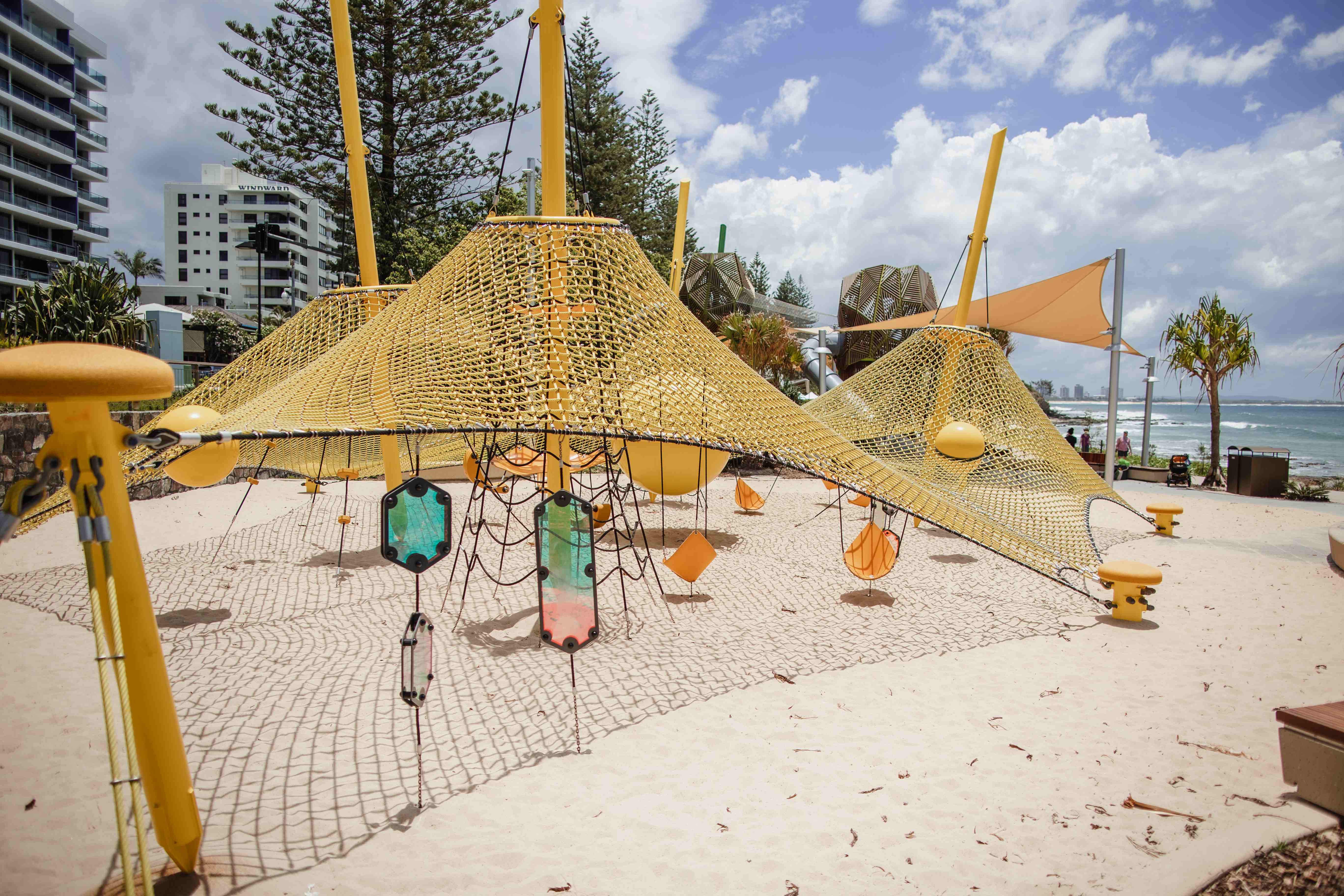 Beachside playground with yellow climbing nets and ropes on sand, modern buildings and ocean visible under a bright cloudy sky