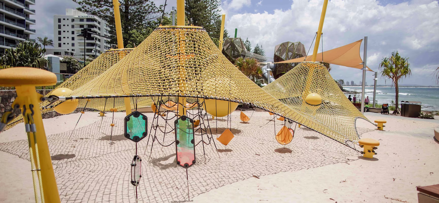 Beachside playground with yellow climbing nets and ropes on sand, modern buildings and ocean visible under a bright cloudy sky