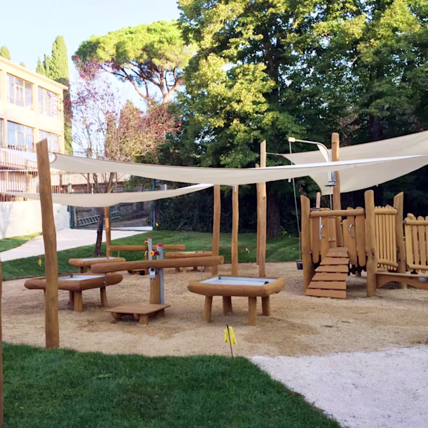 Outdoor wooden playground with benches, sand play tables and shade sails, surrounded by grass and trees near a school building