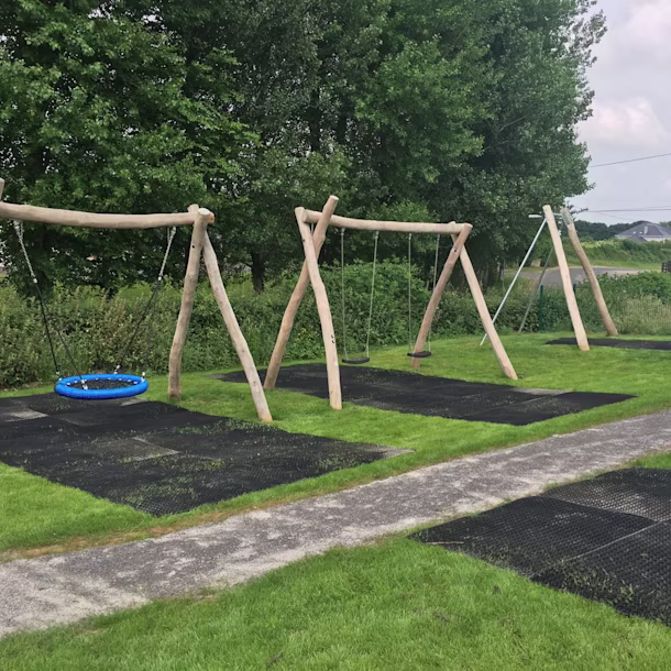 Outdoor wooden playground with benches, sand play tables and shade sails, surrounded by grass and trees near a school building