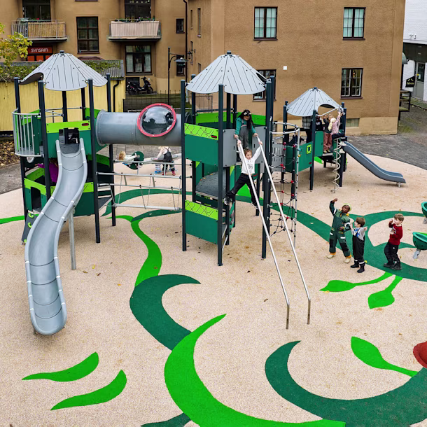 Outdoor wooden playground with benches, sand play tables and shade sails, surrounded by grass and trees near a school building