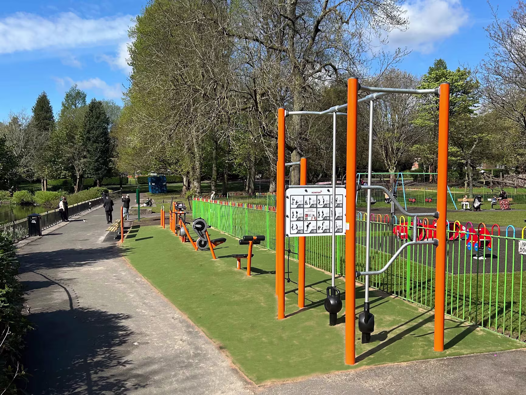 Outdoor park with orange fitness equipment beside a path, trees and blue sky, and a fenced children’s playground with swings in the background
