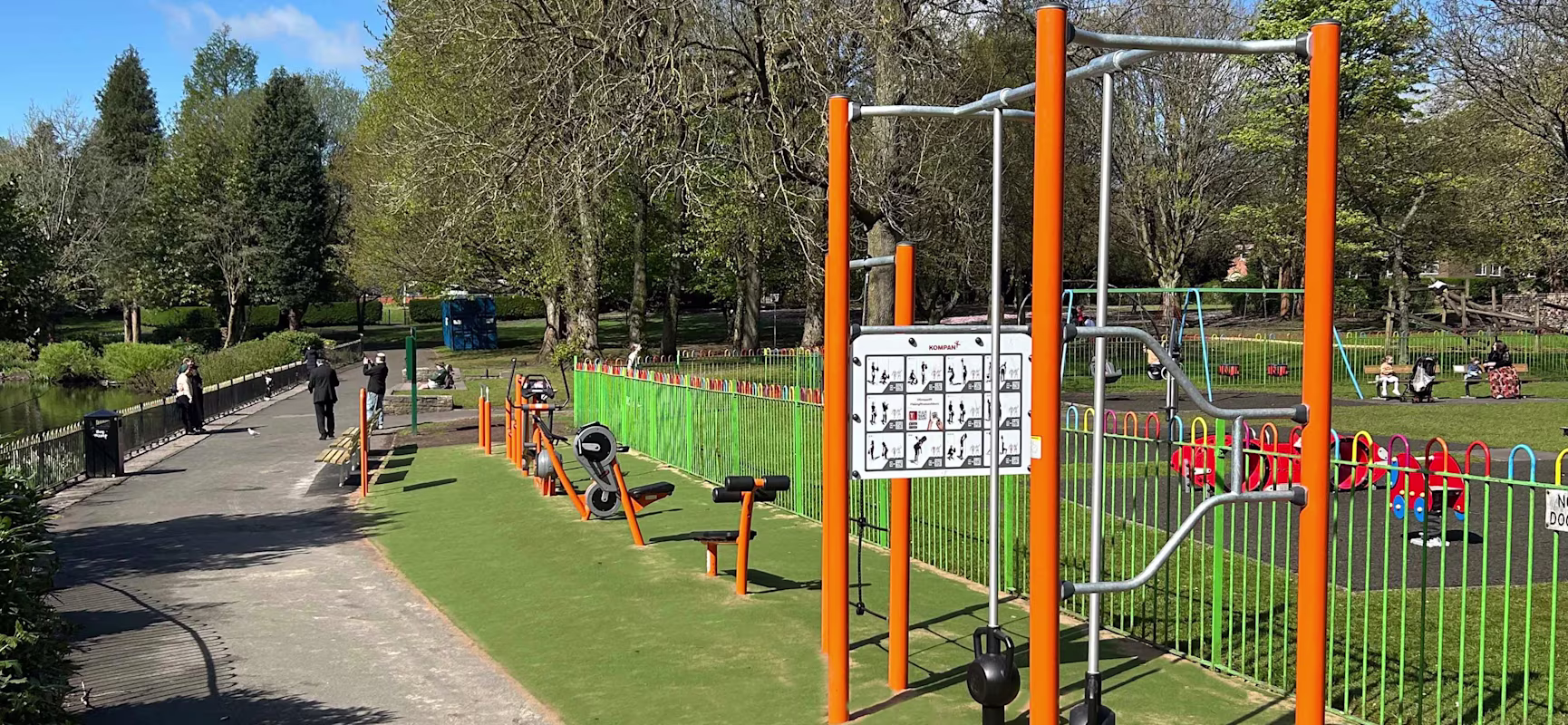 Outdoor park with orange fitness equipment beside a path, trees and blue sky, and a fenced children’s playground with swings in the background