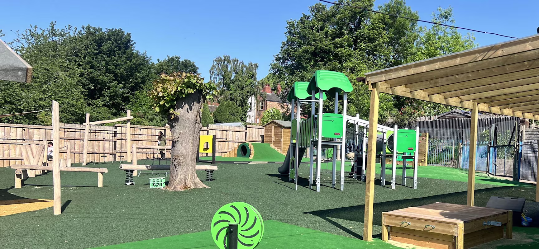 Sunny outdoor playground with green climbing frames, slides, ropes, a tree, and wooden sandpit on bright artificial grass.