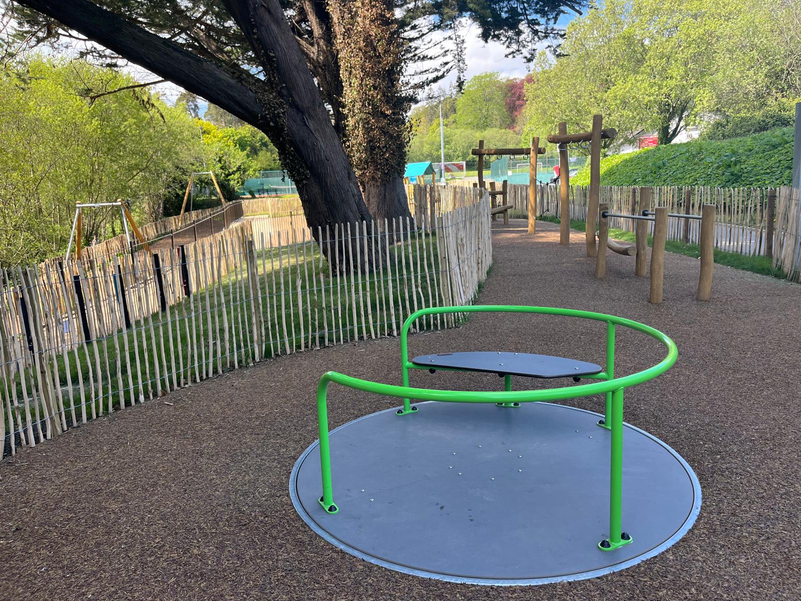 Children playing on a wooden playground structure with slide and climbing frame in a sunny park with trees and buildings nearby