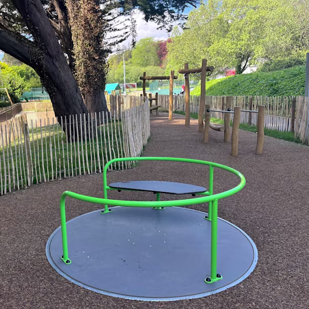 Children playing on a wooden playground structure with slide and climbing frame in a sunny park with trees and buildings nearby