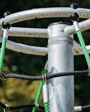 Close-up of a metal playground climbing pole with green and black ropes attached, set against blurred trees in the background