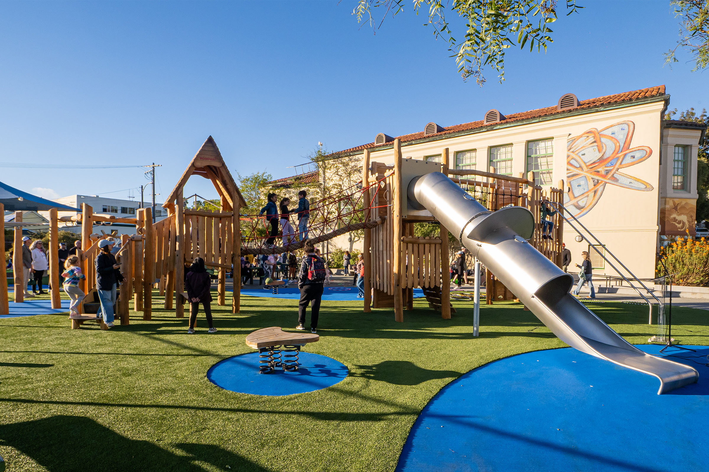 Children play on a wooden playground with rope bridge and tall metal slide on artificial grass, beside a school building with a mural