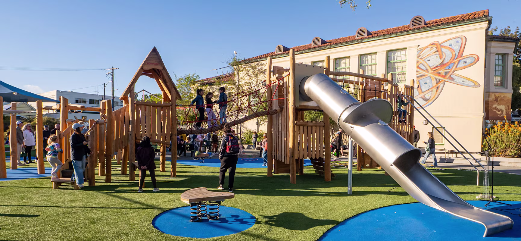 Children play on a wooden playground with rope bridge and tall metal slide on artificial grass, beside a school building with a mural