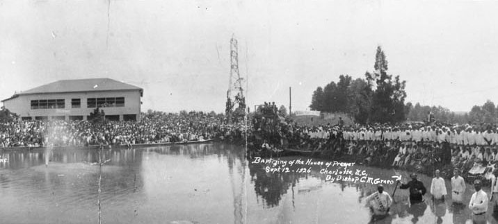 Large crowd gathered beside a pond for an outdoor religious ceremony, with people in white robes standing in the shallow water.