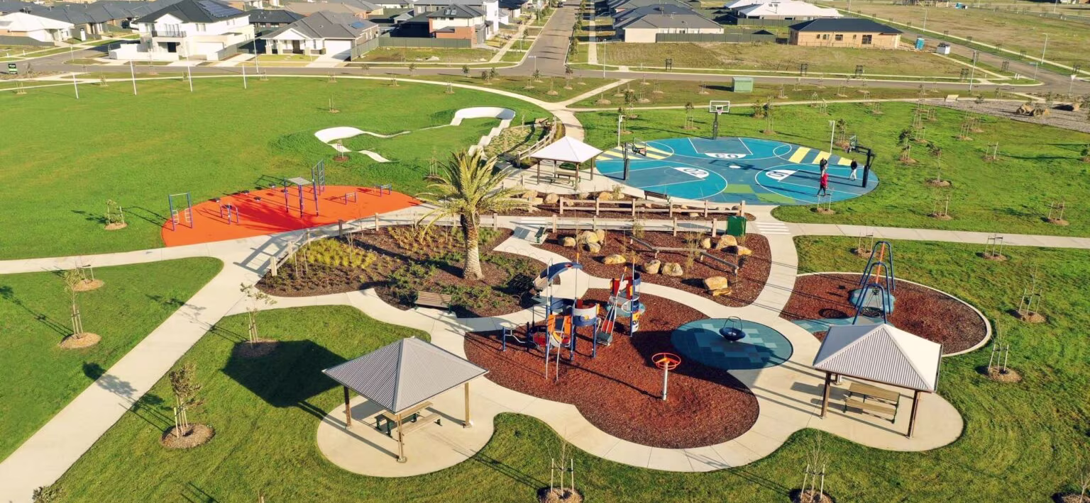 Aerial view of a modern suburban park with playgrounds, basketball court, shelters and paths surrounded by new houses and open fields