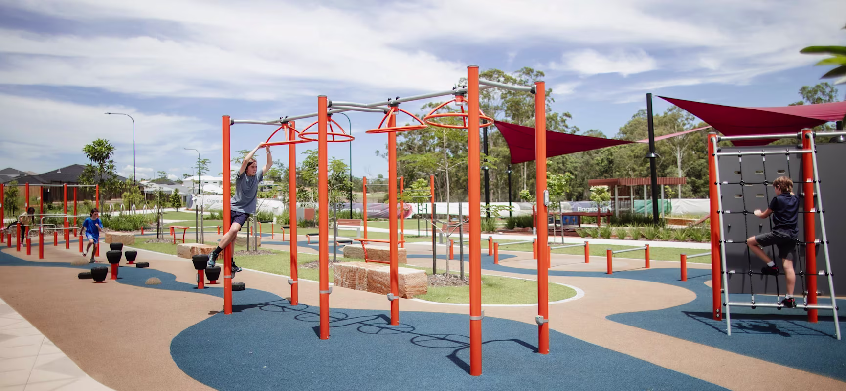 Children play on bright orange climbing equipment in a modern outdoor playground under a blue sky with scattered clouds