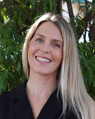 Person with long blond hair in a black sleeveless top standing outdoors in front of green leafy trees, face blurred for privacy