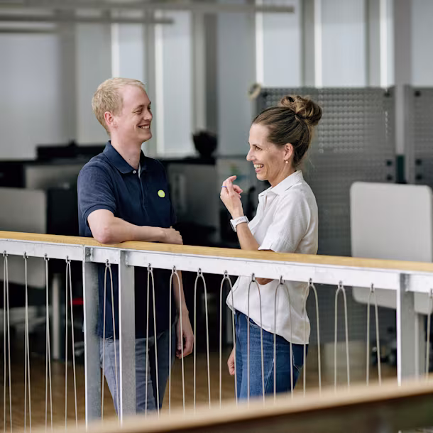 Two people sit in a glass-walled meeting room with laptops on a wooden table, talking under modern circular ceiling lights.