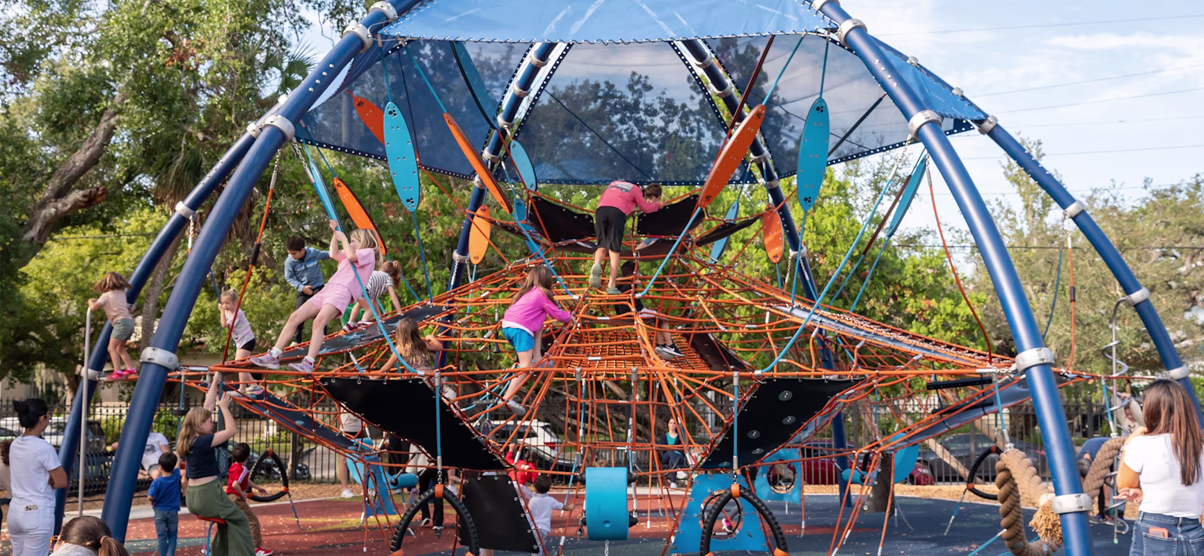 Children climb a large blue and orange rope playground structure under a canopy while adults watch in a sunny park.