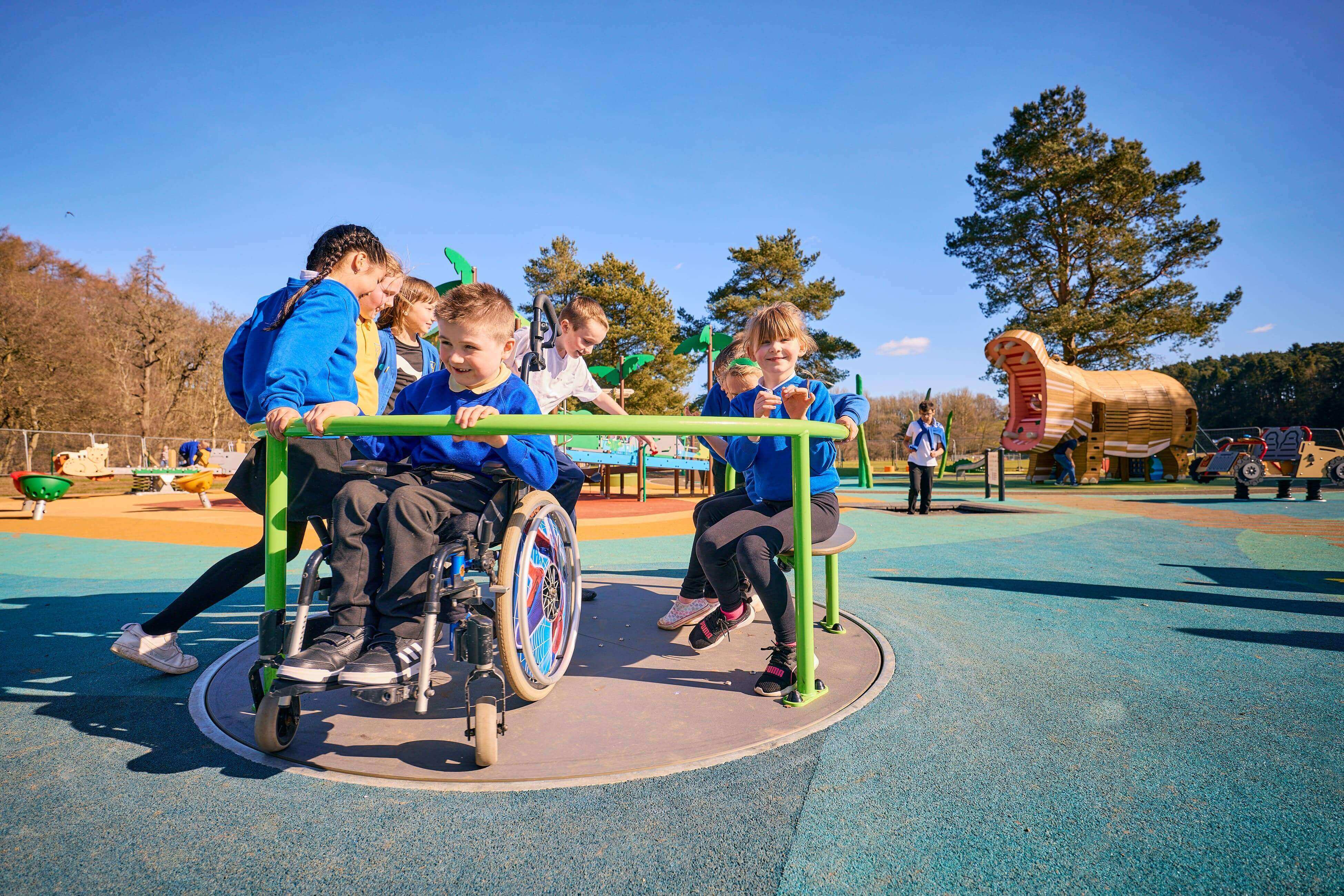 Children play on an inclusive roundabout, one using a wheelchair, in a bright outdoor playground with colourful surfacing and large slides