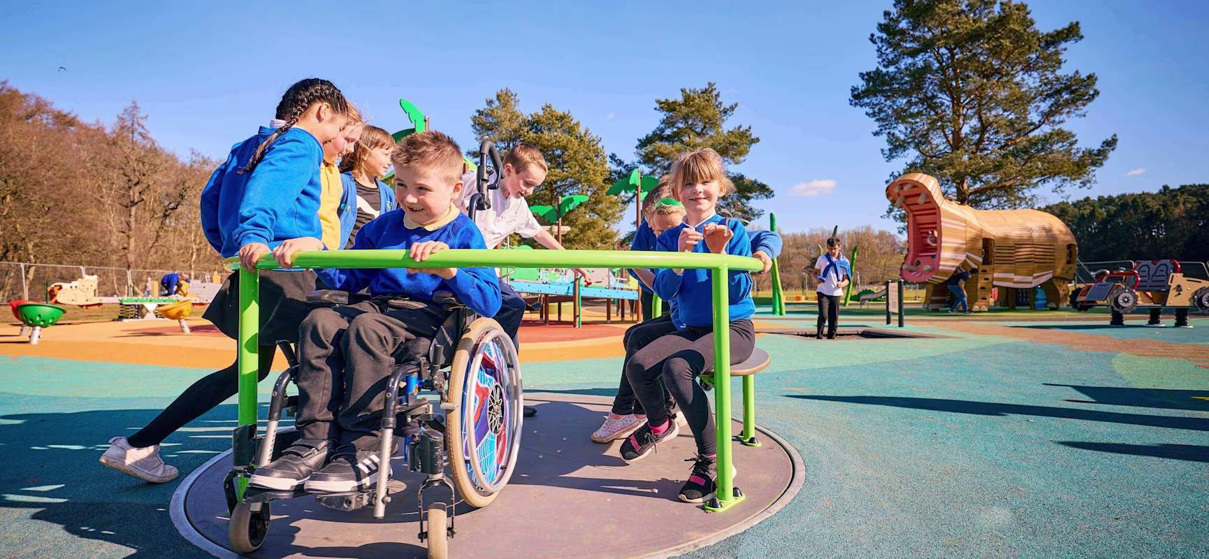 Children play on an inclusive roundabout, one using a wheelchair, in a bright outdoor playground with colourful surfacing and large slides