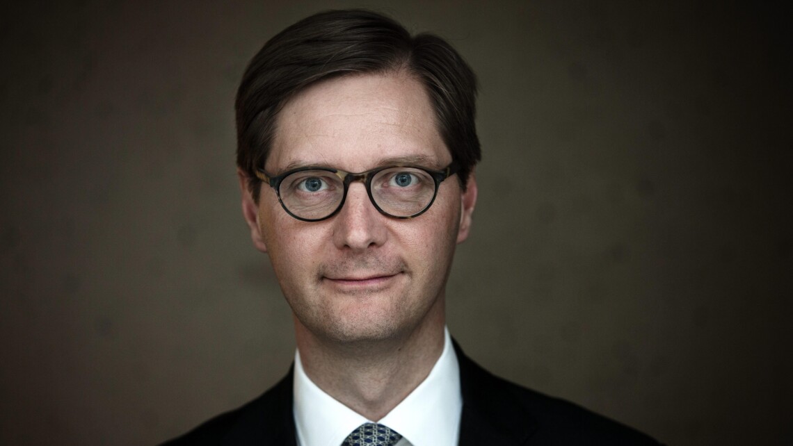 Person in a dark suit and patterned tie, facing forward with their face blurred, against a plain dark background