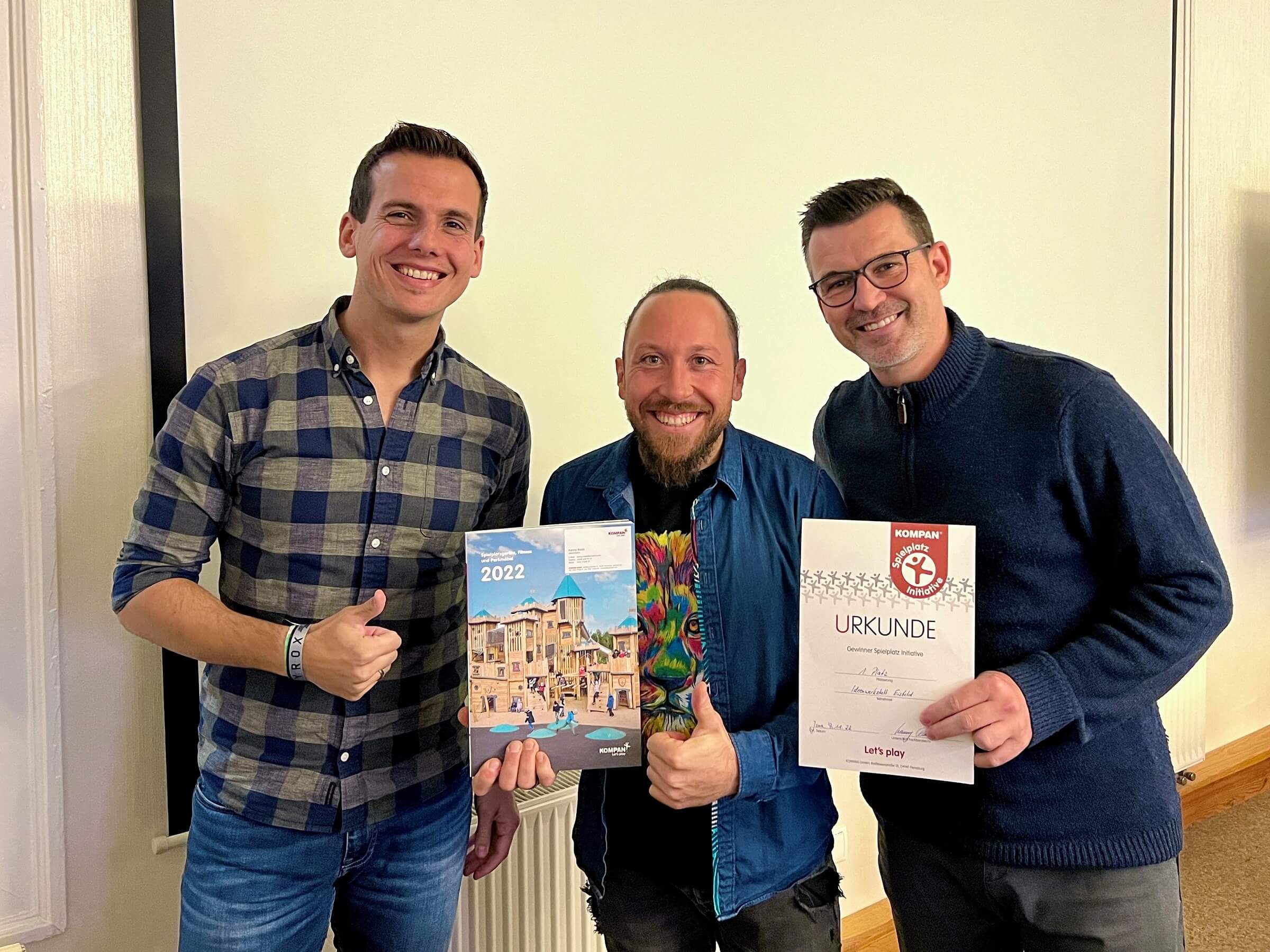 Three people standing indoors, smiling, holding a 2022 catalog and a Kompan certificate, giving thumbs up toward the camera