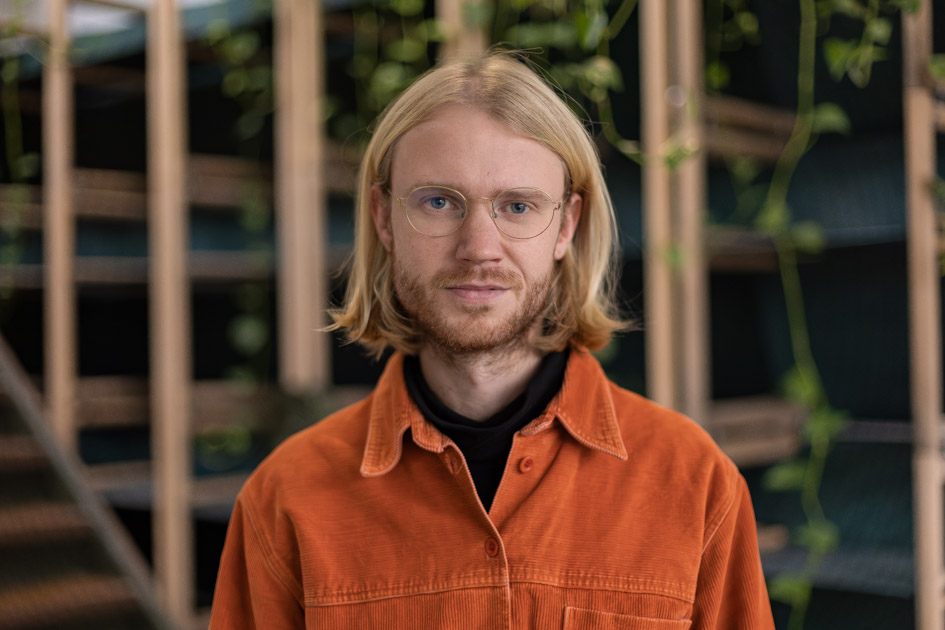 Person with blurred face and long blond hair wearing an orange jacket indoors, with shelves and hanging plants in the background