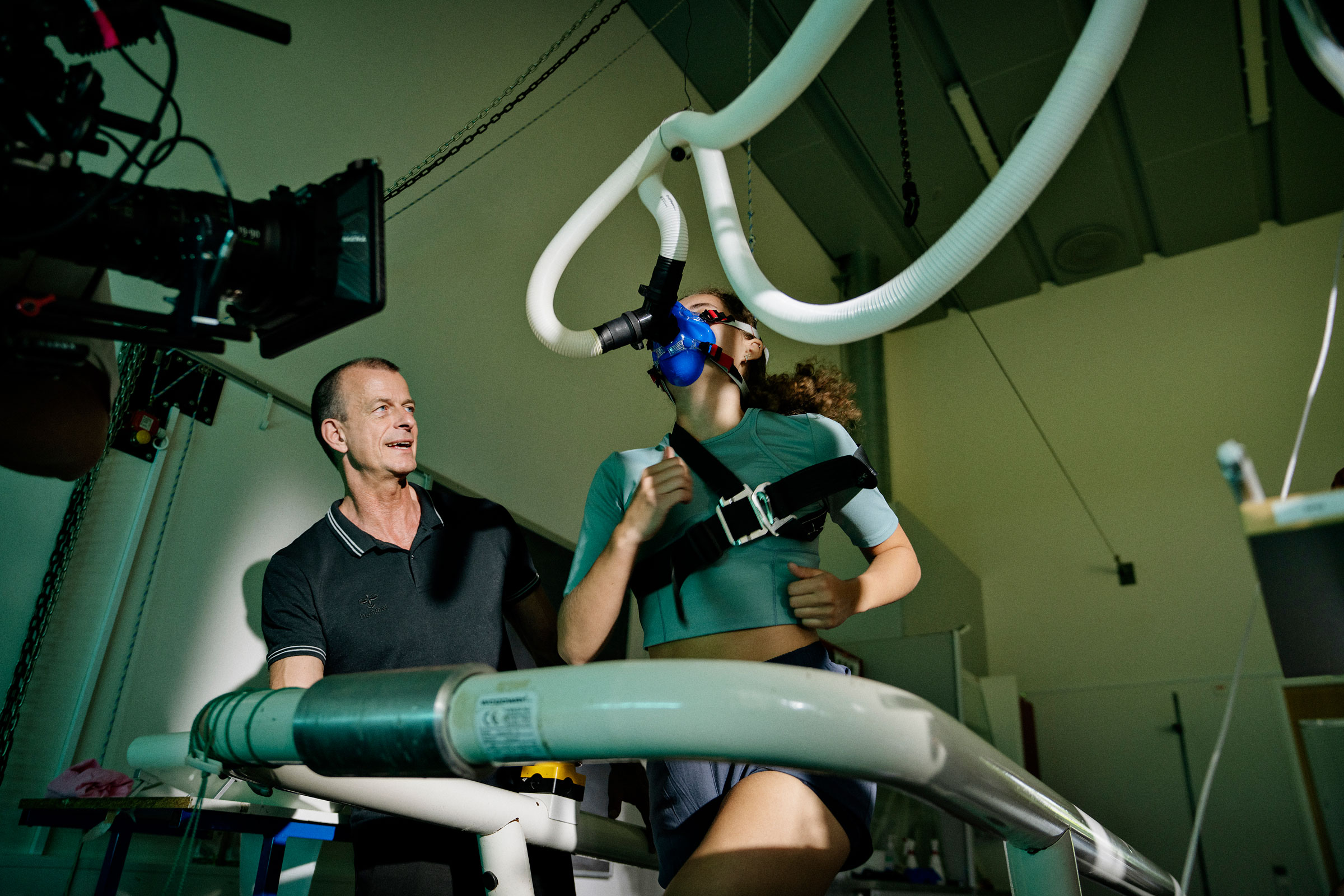 Woman runs on treadmill wearing blue breathing mask and harness while technician supervises in a sports science lab.