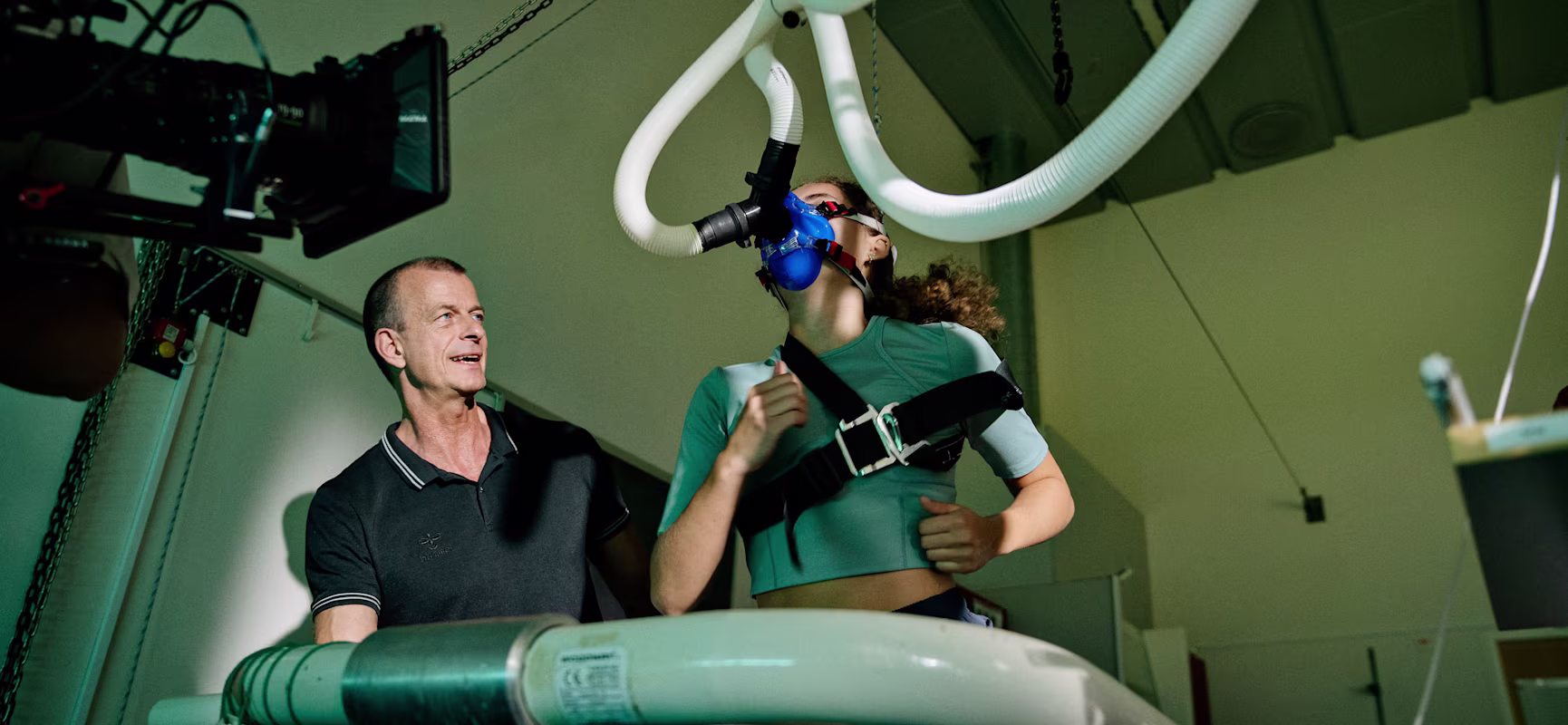 Woman runs on treadmill wearing blue breathing mask and harness while technician supervises in a sports science lab.