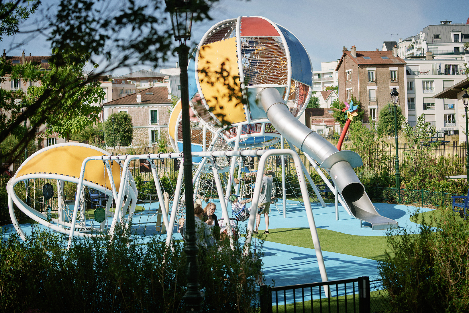 Children play on a large colourful metal playground with slides and climbing nets in a sunny urban park surrounded by greenery and flats