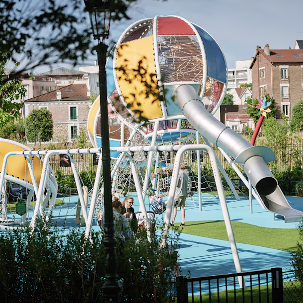 Children play on a large colourful metal playground with slides and climbing nets in a sunny urban park surrounded by greenery and flats
