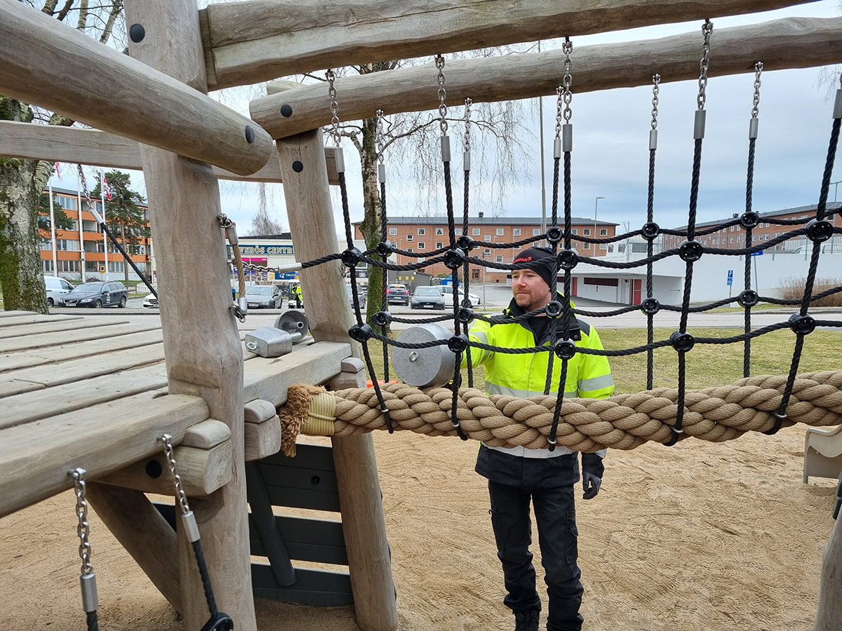 Worker in high-vis jacket stands on sandy playground, inspecting a rope bridge and climbing net with buildings and cars in background