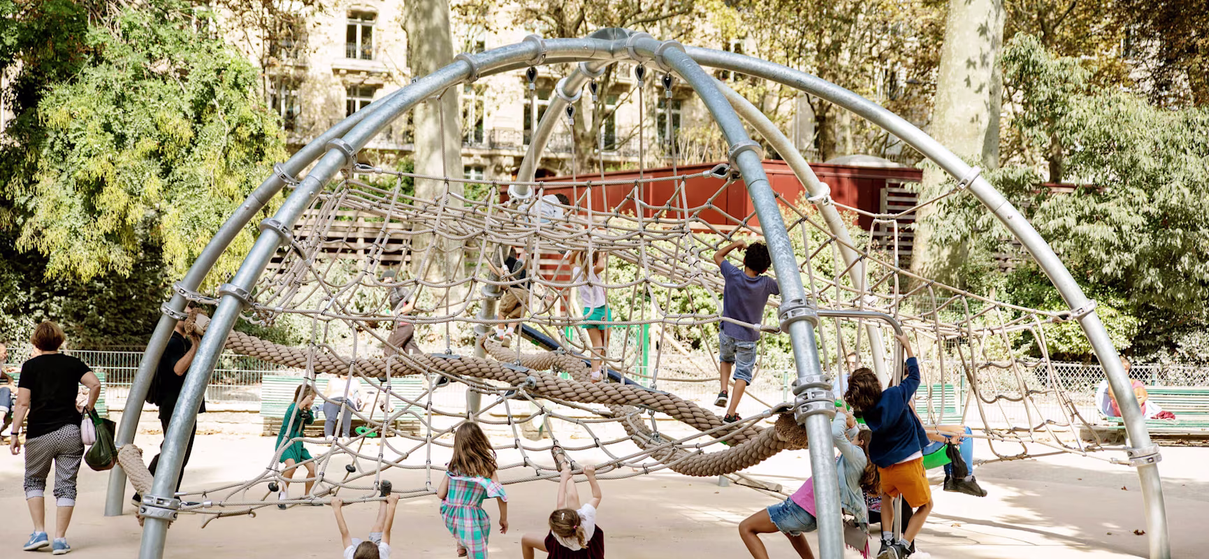 Children climb and swing on a large rope and metal dome playground structure in a sunny park with trees and buildings behind