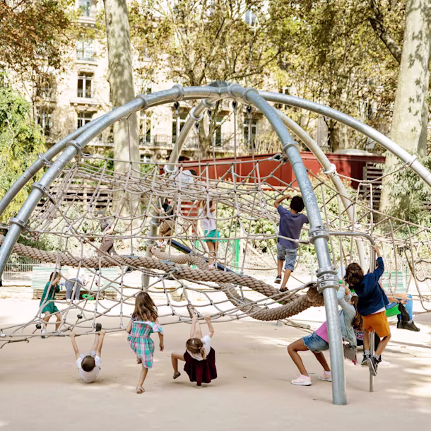 Children climb and swing on a large rope and metal dome playground structure in a sunny park with trees and buildings behind