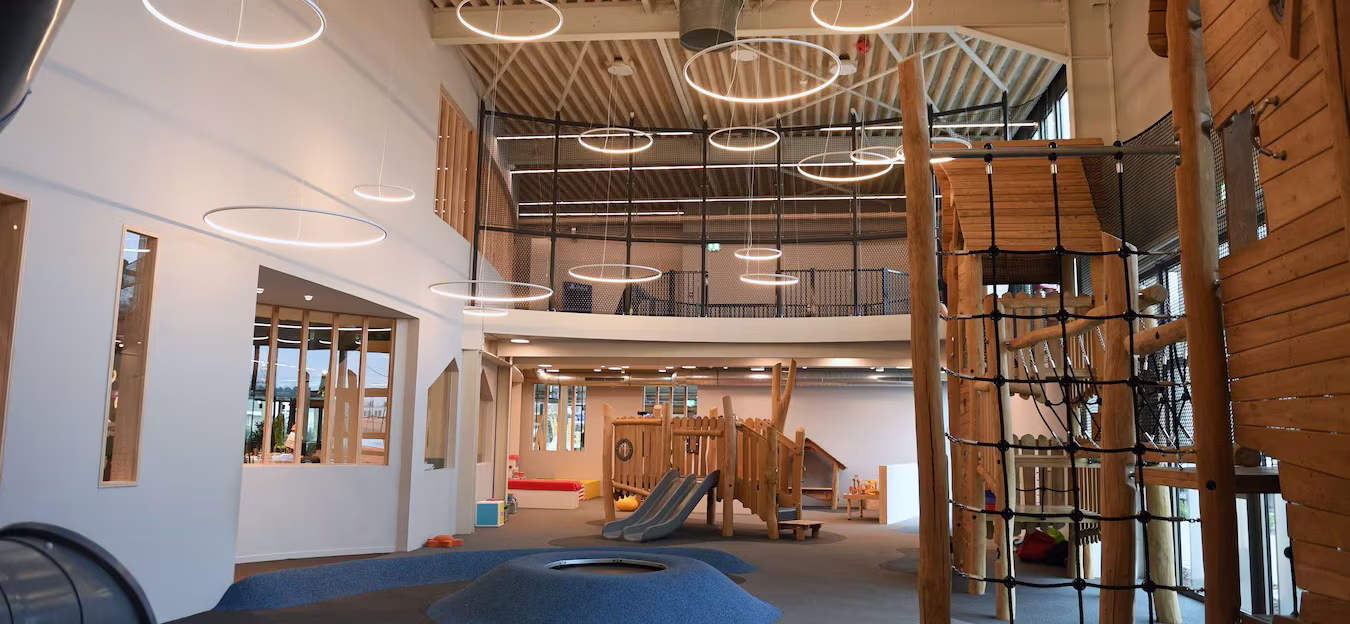 Modern indoor playground with wooden climbing structures, nets, blue rubber hills and circular ceiling lights in a spacious room.